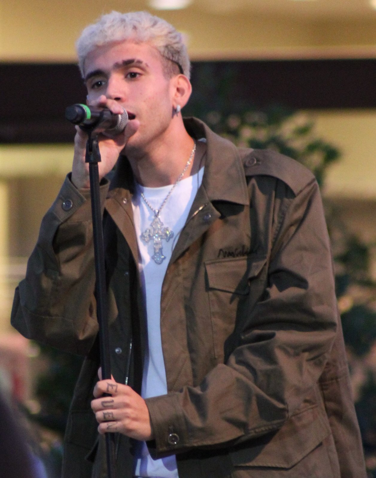 Aka 7even canta al microfono su un palco, capelli biondo platino e giacca militare