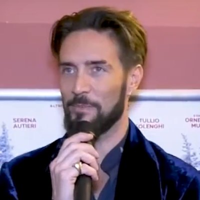 Alex Belli parla al microfono durante una conferenza stampa, giacca blu scura