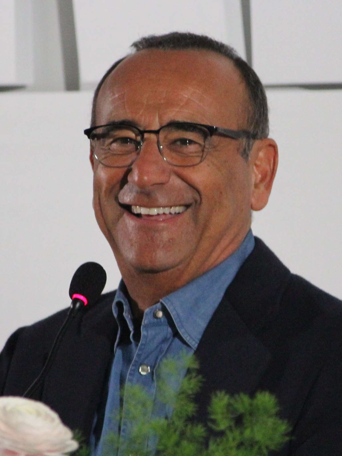 Carlo Conti sorride in primo piano, occhiali scuri e camicia di jeans