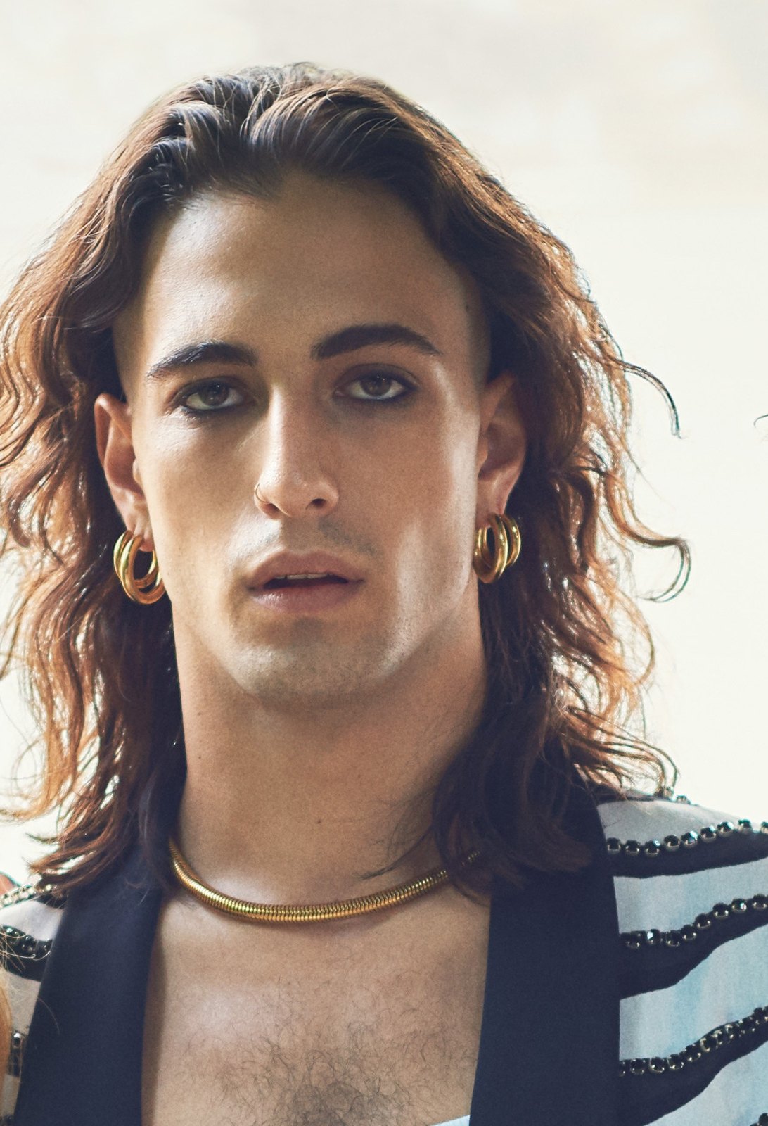 Damiano David dei Måneskin guarda dritto in camera, capelli lunghi e orecchini dorati