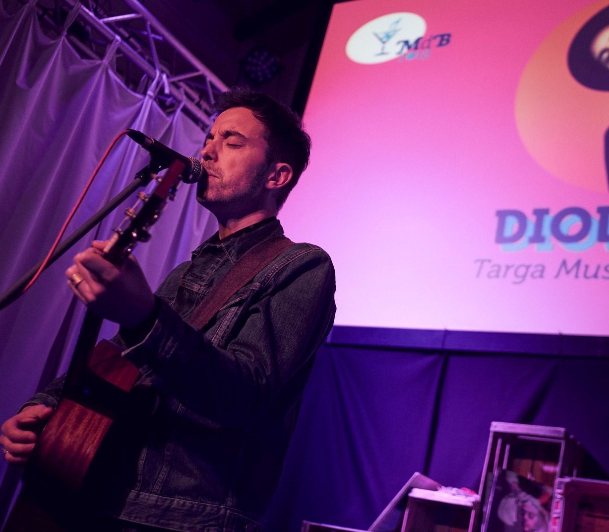 Diodato canta al microfono con la chitarra su un palco illuminato di viola