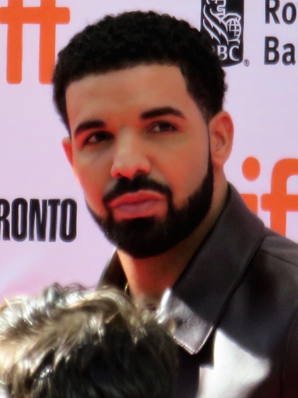 Drake, barba curata e giacca scura, guarda di lato su un red carpet