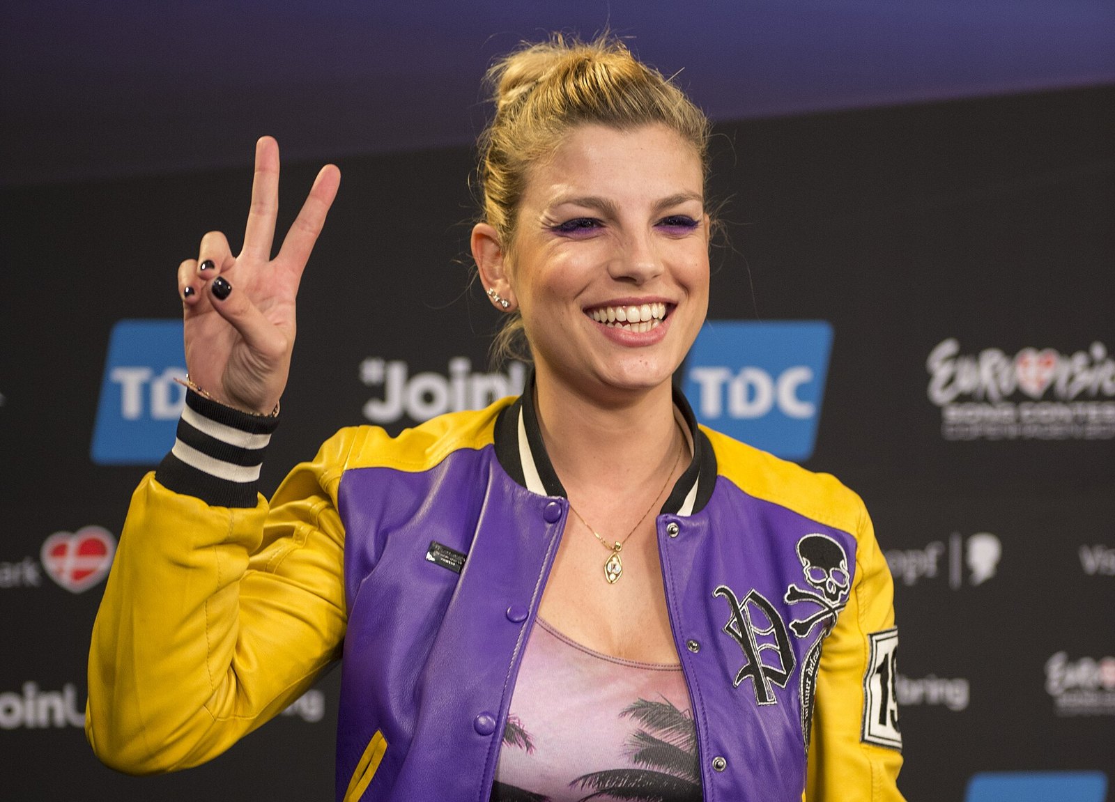 Emma Marrone sorride e fa il segno della vittoria con le dita davanti a un fondale