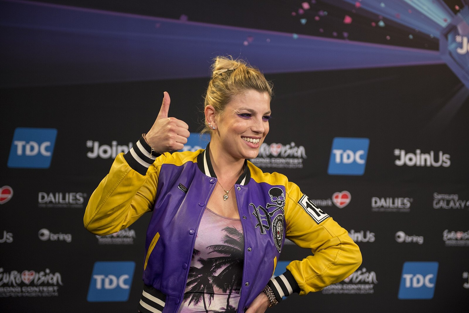 Emma Marrone sorride e fa il pollice in su davanti a un pannello di sponsor