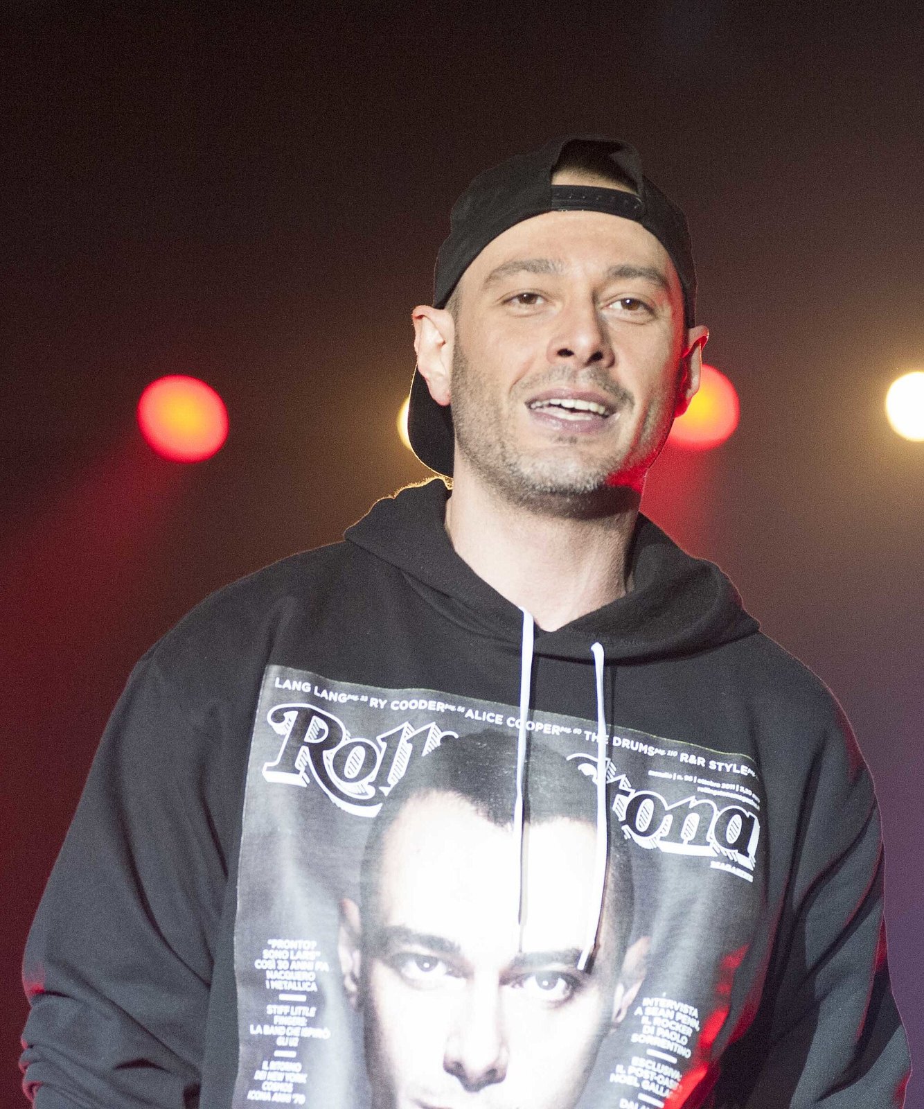 Fabri Fibra sul palco con il cappellino girato al contrario, guarda verso il pubblico