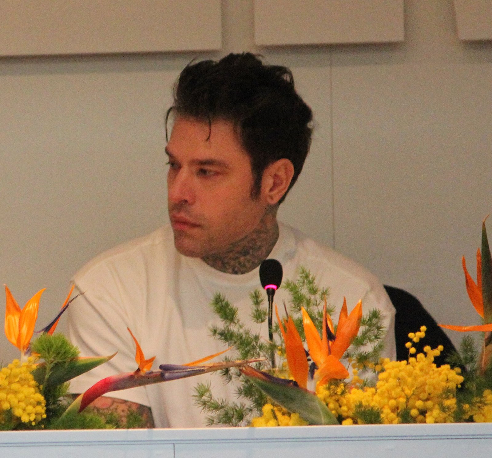 Fedez seduto al tavolo di una conferenza stampa, maglietta bianca, mentre guarda di lato