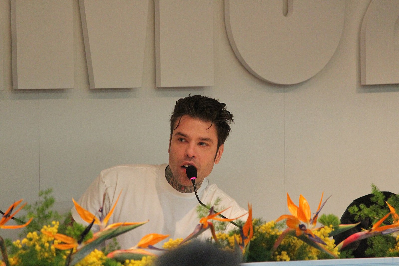Fedez parla al microfono in conferenza stampa, maglietta bianca e sfondo chiaro