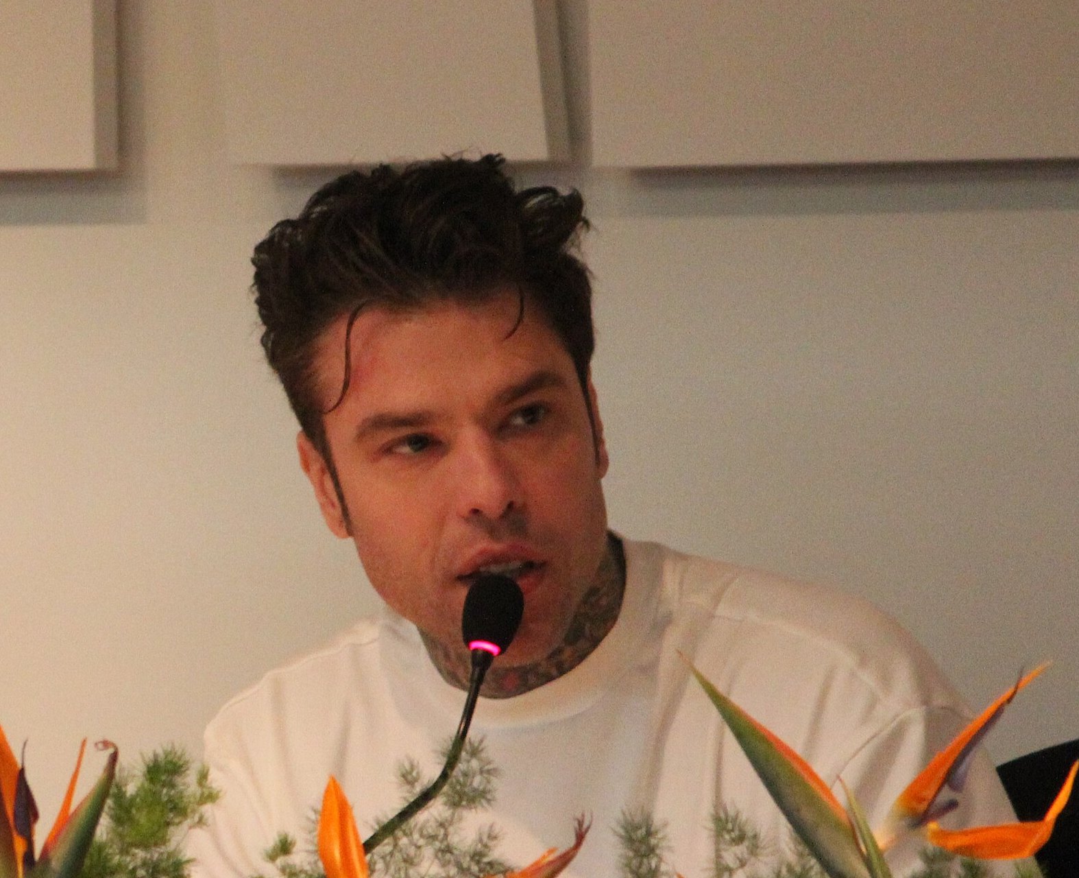Fedez parla al microfono in conferenza stampa a Sanremo 2026, maglietta bianca e tatuaggi sul collo