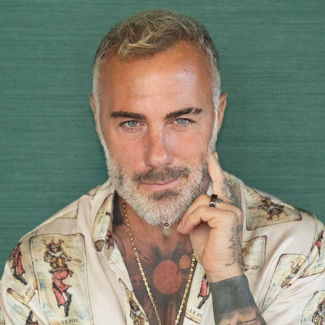 Gianluca Vacchi guarda in camera con la mano appoggiata al viso, barba bianca e camicia fantasia