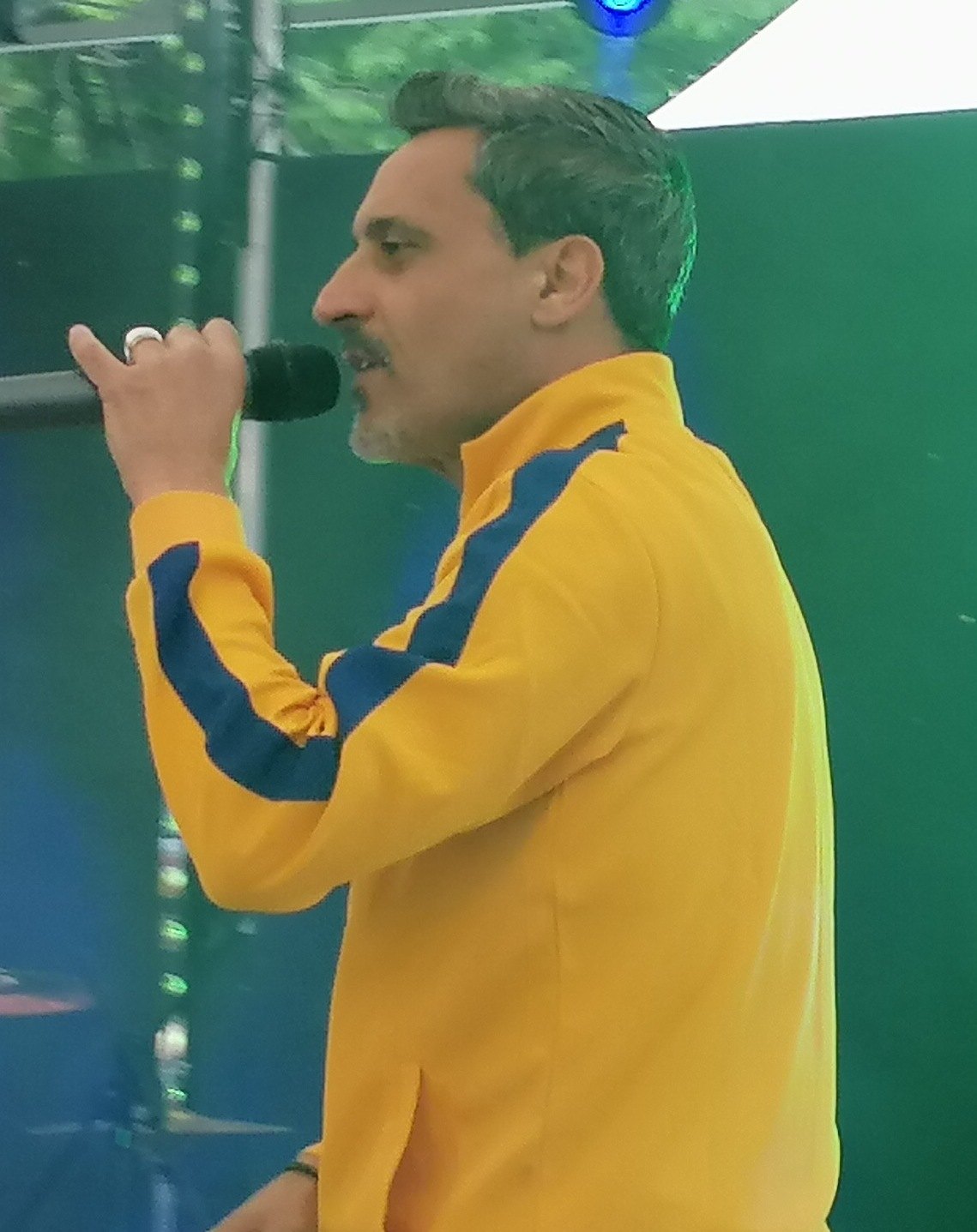 Giorgio Vanni canta al microfono su un palco, con una giacca gialla e blu