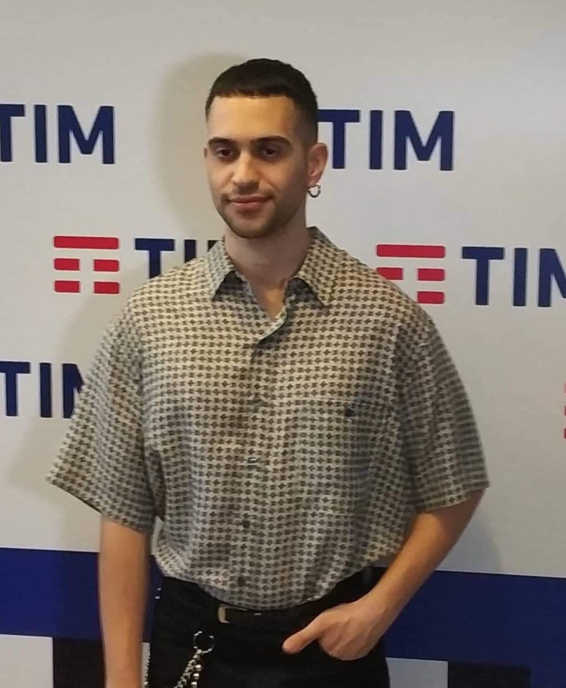 Mahmood in camicia a fantasia davanti a un muro con i loghi di un evento