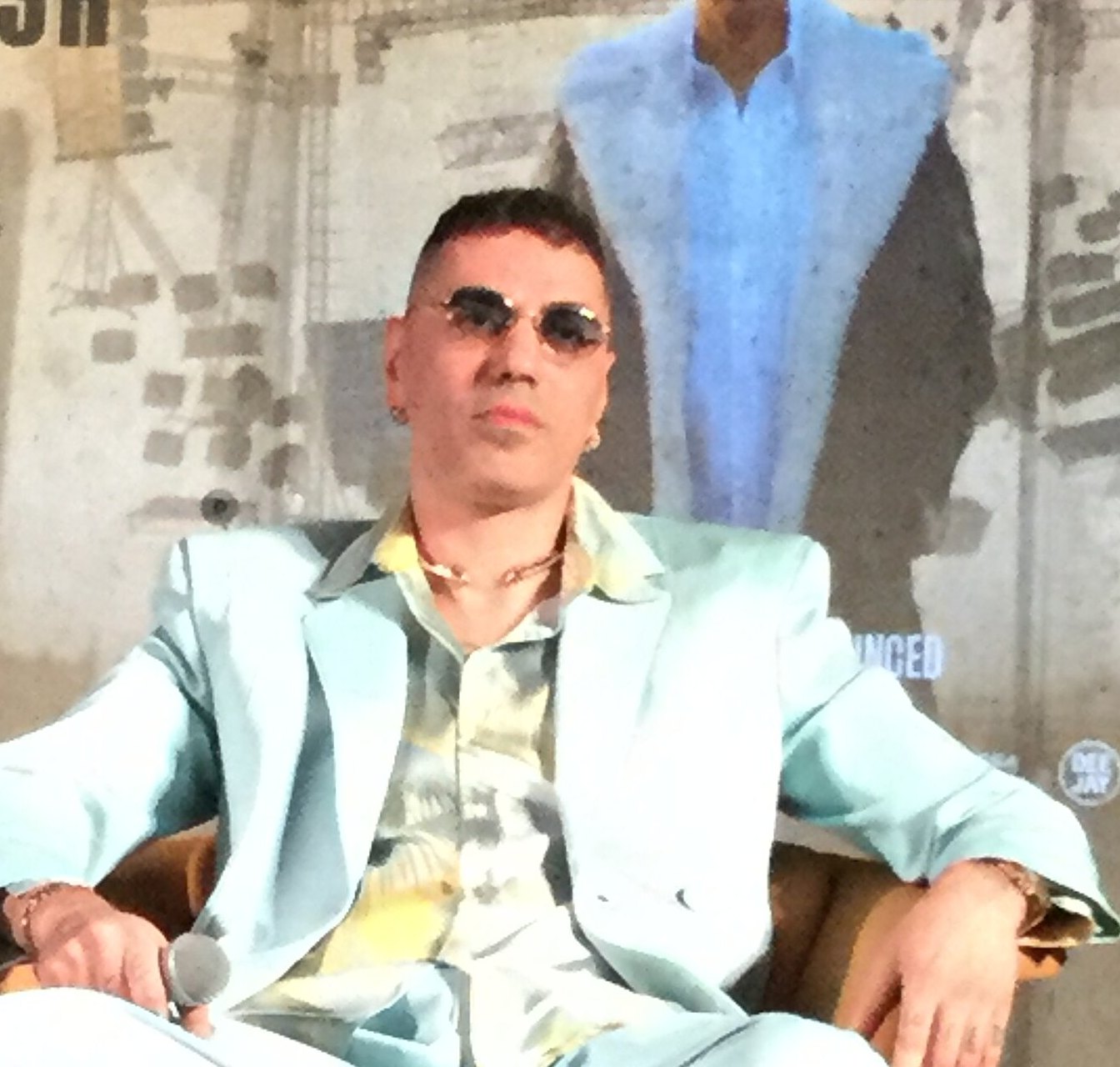 Marracash seduto con occhiali da sole davanti al manifesto del Marrageddon