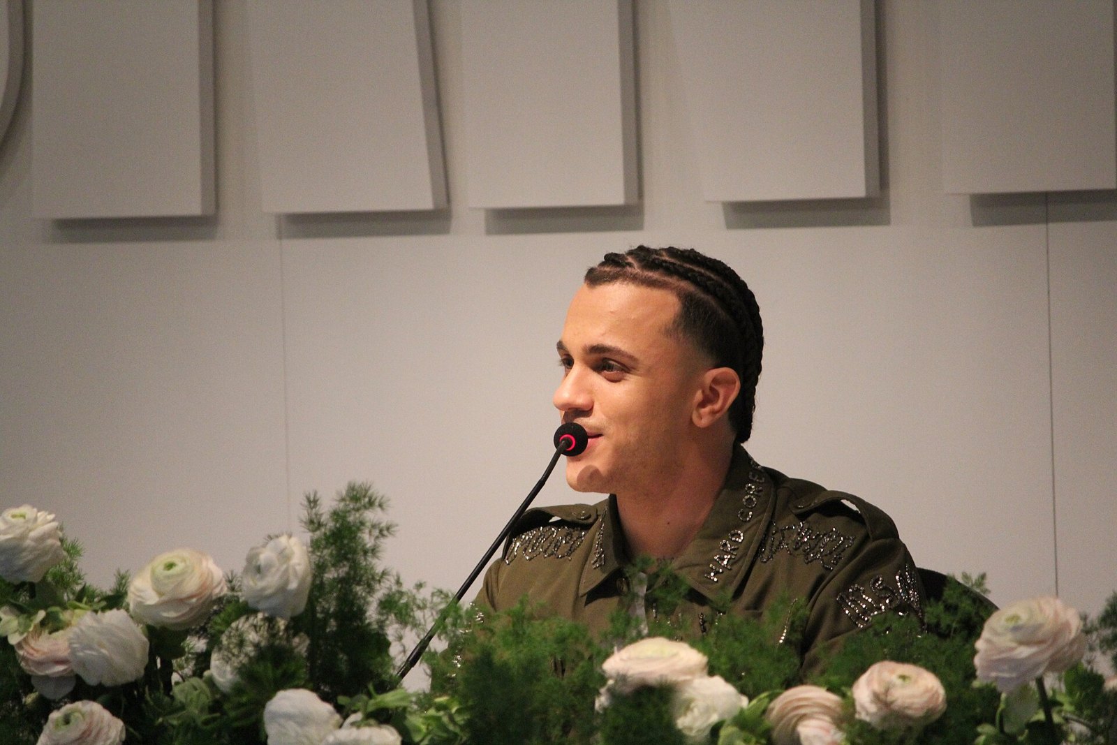 Samurai Jay parla al microfono durante una conferenza stampa, seduto dietro a dei fiori bianchi