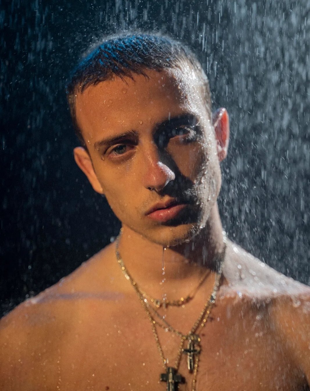 Sangiovanni in primo piano sotto un getto d'acqua, con i capelli corti e lo sguardo dritto in camera